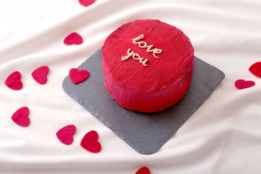 A small delicious red bento cake with the inscription -I love you, next to which red hearts are scattered on a white bed.To close up. Copy space