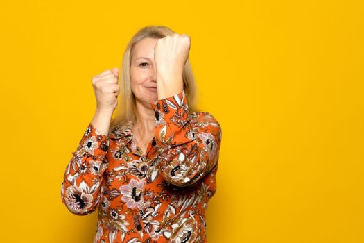 Beautiful blonde woman on vacation in patterned dress on yellow background. Ready to fight with fist defense gesture, confident smiling face, not afraid of trouble