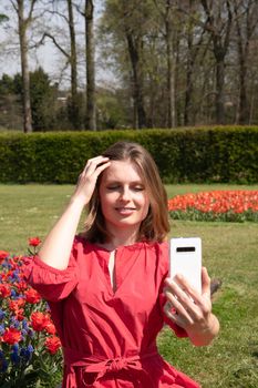 A young beautiful girl in a red dress in a spring park plays with her hair, flirts and takes a selfie on a mobile phone, tulips bloom and people walk in the park, pamper themselves.High quality photo