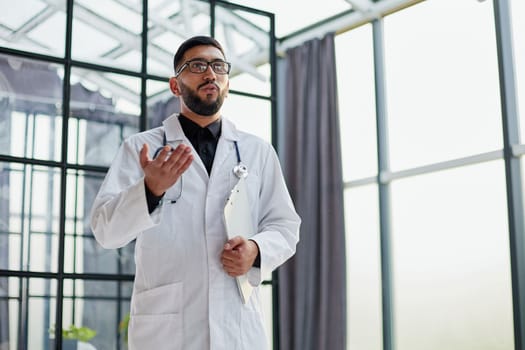 Portrait of cheerful joyful doc with bristle in white lab coat, tie and stethoscope on his neck, having clipboard in hands,