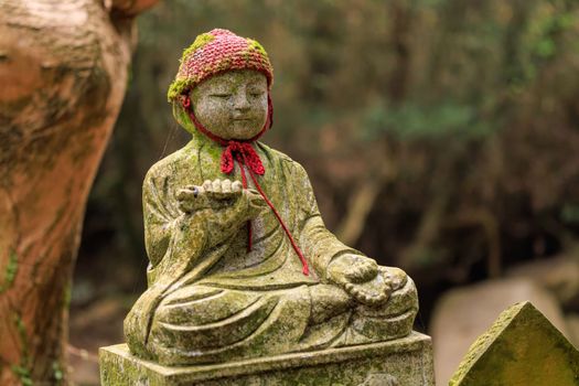 Stone statue of child sitting cross-legged wearing a knit cap at Japanese shrine in woods. High quality photo