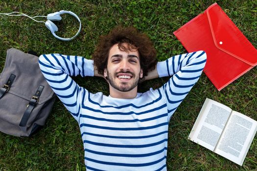 Top view of happy, smiling young caucasian man relaxing lying down on grass looking at camera. Education and campus concept.