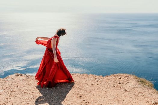 Side view a Young beautiful sensual woman in a red long dress posing on a rock high above the sea during sunrise. Girl on the nature on blue sky background. Fashion photo.