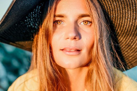 Portrait of happy young woman wearing summer black hat with large brim at beach on sunset. Closeup face of attractive girl with black straw hat. Happy young woman smiling and looking at camera at sea