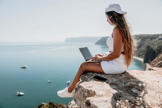 Successful business woman in yellow hat working on laptop by the sea. Pretty lady typing on computer at summer day outdoors. Freelance, travel and holidays concept.