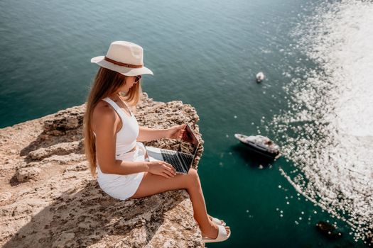 Successful business woman in yellow hat working on laptop by the sea. Pretty lady typing on computer at summer day outdoors. Freelance, travel and holidays concept.