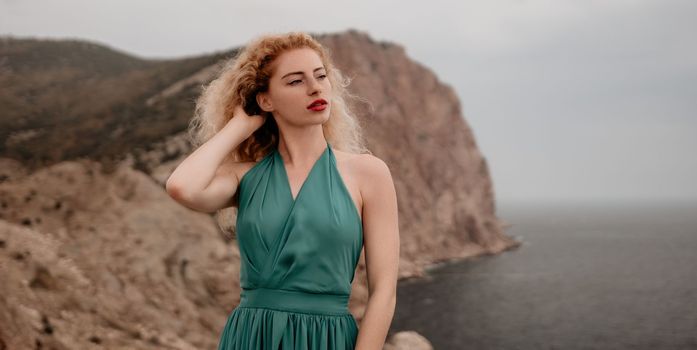 Side view a Young beautiful sensual woman in a mint long dress posing on a volcanic rock high above the sea during sunset. Girl on the nature on overcast sky background. Fashion photo