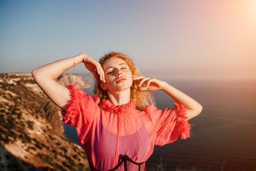 Close up shot of beautiful young caucasian woman with curly blond hair and freckles looking at camera and smiling. Cute woman portrait in a pink long dress posing on a volcanic rock high above the sea
