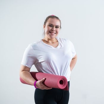 Young fat caucasian woman holding a sport mat. Charming plus size model in sportswear stands on a white background.