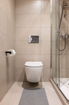 Toilet with walls and floor in soft beige square tiles. The white toilet creates the effect of floating above the floor. The shower cabin is closed by sliding glass doors and protects from splashes