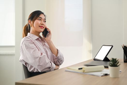 Confident female investment advisor talking on mobile phone with client and using digital tablet at her workstation.