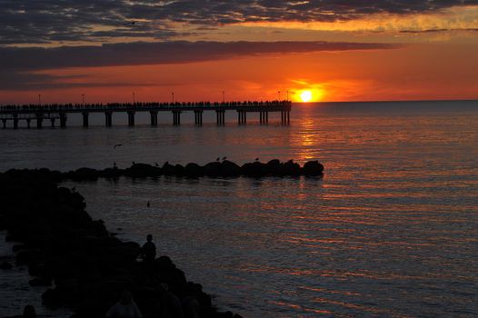 people silhouette standing on wooden pier at the sea with beautiful bloody sunset. A long exposure golden sunset view of a wooden jetty along the coast with calm sea