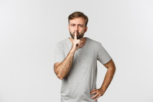 Angry bearded man in grey t-shirt, hushing and frowning, scolding person for talking too loud, standing over white background.