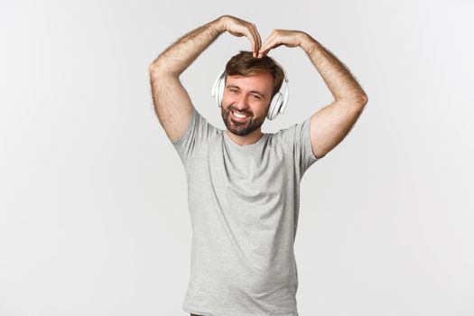 Cute man with beard, showing big heart and listening music in headphones, smiling silly at camera, standing over white background.