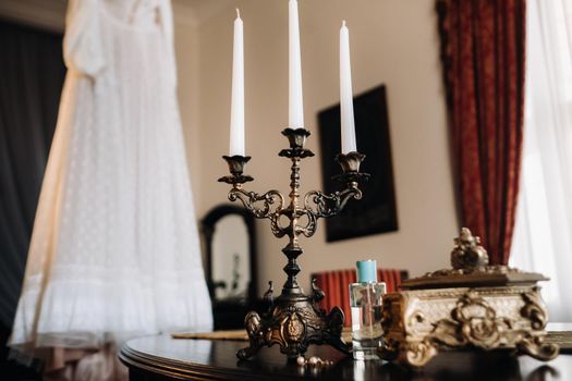 Vintage wedding dress hanging on a wooden hanger and a candle holder standing on a table in the castle.