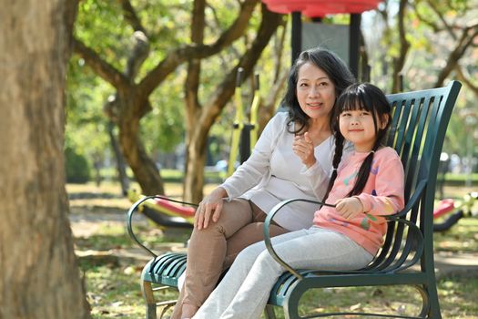 Image of cute little grandchild and elderly grandmother having casual conversation, while sitting on a bench in the park.