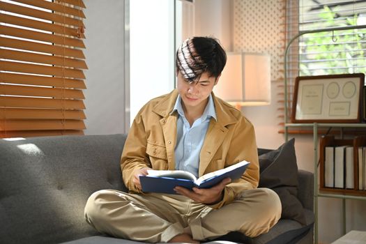 Peaceful millennial asian man reading book while resting on couch in bright living room interior.