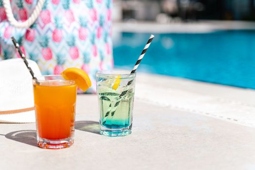 Tropical sparkling lemonade cocktails by the pool with pink beach bag and white hat in the background. Picture of glasses with orange and mint lemon fruit cocktails. Hello summer holiday vacation.