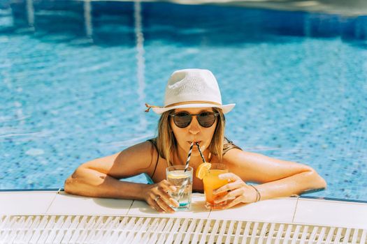 Portrait of beautiful cute smiling young woman lady girl in a bikini, hat, and sunglasses holding tropical lemonade cocktail while chilling in swimming pool. Hello summer holiday vacation