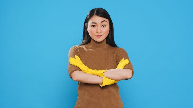 Asian professional maid smiling at camera after finishing house cleaning, standing in studio over blue background. Housekepper was skilled in the art of housekeeping, ensuring every surface was clean