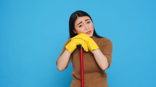 Tired housekepper falling asleep on broom while cleaning client home, being overworked in studio over blue background. Sleepy maid wearing rubber gloves while being was responsible for the houseclean