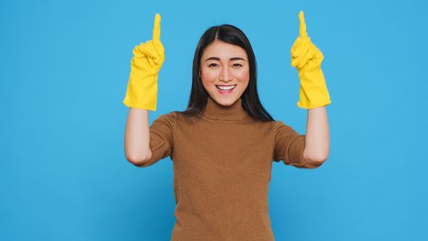 Professional housekepper pointing and showing cleaning advertisement or isolated text, standing on blue background in studio. Housewife worked hard to maintain a clean home, using hygiene products