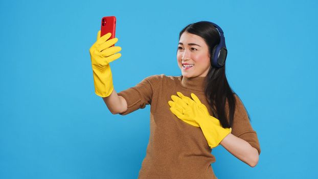 Smiling asian maid wearing headphone while talking with remote friend during videocall meeting on phone, standing in studio over blue background. Housekeeper doing house cleaning using chemical spray