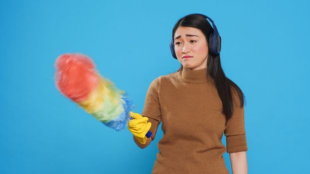 Exhausted sleepy housekeeper wearing headphones while doing cleaning in house using feather duster, yawning in front of camera. Overwhelmed maid role was vital to the cleanliness of many homes