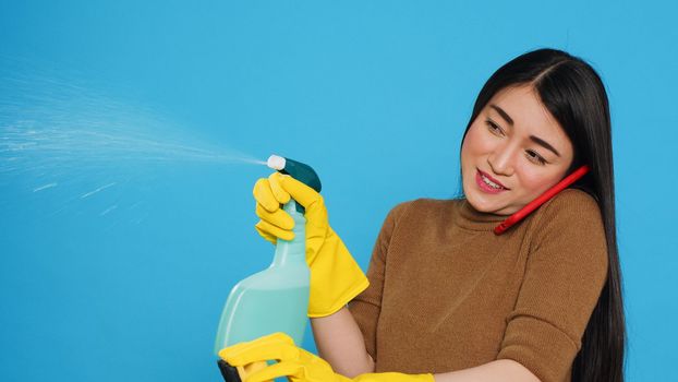 Asian housekeeper using duster and protective gloves to clean house while talking at smartphone with remote friends, having remote discussion. Housekeeping and cleaning concept.