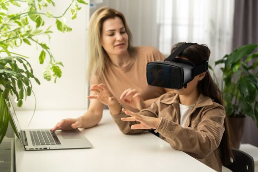 Blond excited mother helping her adorable girl teenager virtual using virtual reality headset at home interior