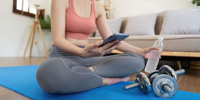 Woman sit on mat holding phone watching online workout training tv class video, using fitness app at home.