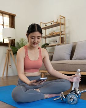 Woman sit on mat holding phone watching online workout training tv class video, using fitness app at home.