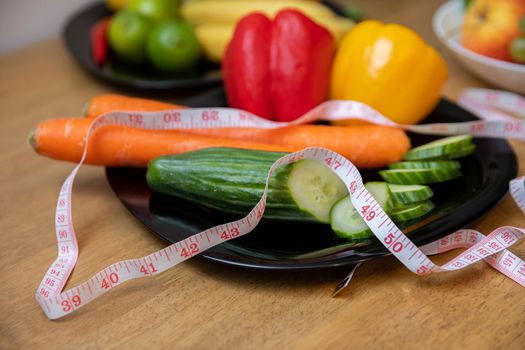 Close-up of a black plate full of vegetables such as cucumber, peppers, carrots. Measure tape lies on top of the vegetables. In the background, a smudged plate of fruit.