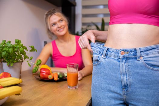 In the foreground, a close-up on the torso of a woman standing forward and pulling back her loose jeans from the side. In the background, a girl looking at her friend. Next to a table set with fruits and vegetables
