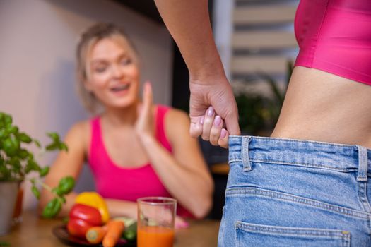 A girl standing with her back shows her friend sitting in the background the effects of weight loss by pulling back too loose pants at the waist. Women wear matching pink sports tops