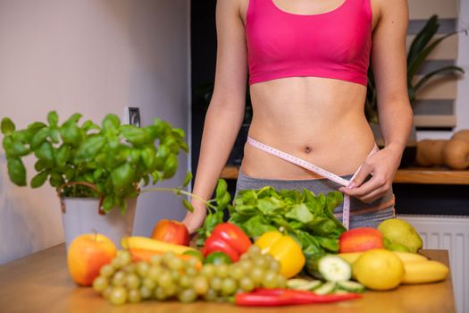 an athletic woman with a narrow waist wraps measure tape around her waist. In front of the woman's figure stands a table and on it lie colorful vegetables, fruits and herbs. In the background is a window and a windowsill with a pumpkin.