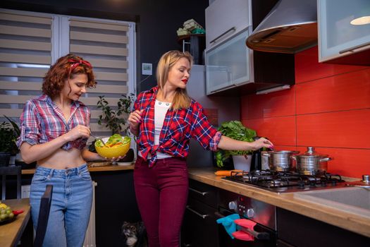 A woman with curly hair holds a yellow bowl and stirs in it. A blonde woman standing by the stove stirs in a metal pot and smiles. Both are dressed in plaid shirts. A dog sits on the floor.