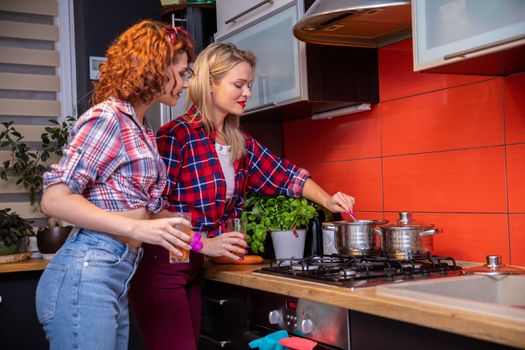 Two women cooking dinner in the kitchen lean over a gas stove and look into a metal pot, one of them stirring in the pot with a spoon. Both are dressed in checkered shirts.