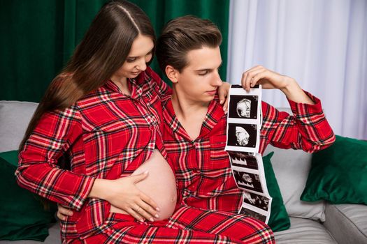 A man and a woman waiting to become parents sit on a gray couch. The woman has an exposed pregnant belly. The man is holding printouts of ultrasound images.