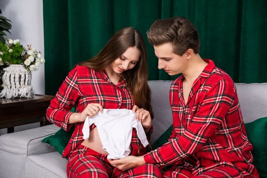 An expecting couple try on infant sleepwear for the woman's exposed pregnant belly. The parents-to-be sit together on a gray couch and are wearing identical red plaid pajamas.