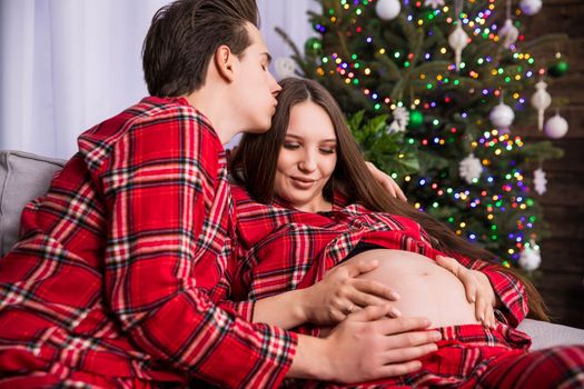 A couple sitting near a Christmas tree visible in the blurry background is expecting a baby. The woman is advanced in her pregnancy, and the man is kissing her on the top of her head.