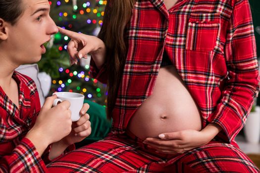 The tip of a man's nose, seen in profile, is touched by a woman sitting next to him. The woman is pregnant, a close-up of a large pregnant belly.