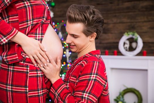 Kneeling man. Close-up of the pregnant belly in profile. The man kneels in front of his pregnant wife and holds his hand on her belly. The couple is dressed in identical red pajamas.