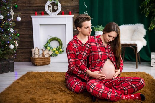A man embraces a woman while sitting behind her back. The woman is advanced in pregnancy and exposes her exposed belly. Behind the couple in the background are Christmas decorations and a Christmas tree.