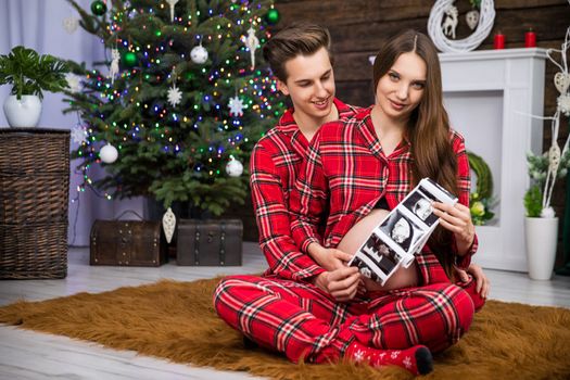 A man sits behind a woman on a carpet and embraces her pregnant belly. The woman shows an ultrasound photo of the baby. The man is smiling and the woman is looking straight into the lens.