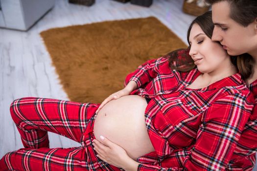 A pregnant woman has her hands on her stomach. A husband embraces his wife. The man kisses his partner on the cheek. A Christmas tree, a potted plant and a brown carpet can be seen in the background.