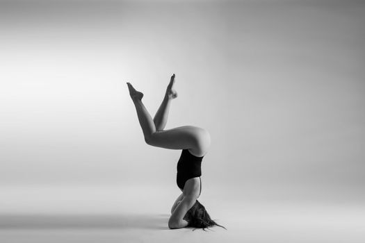 Young slim female in black bodysuit makes yoga stretching exercise on white studio background.