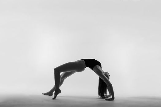 Young slim female in black bodysuit makes yoga stretching exercise on white studio background.