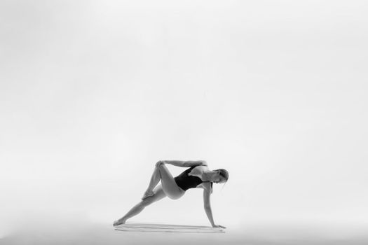 Young slim female in black bodysuit makes yoga stretching exercise on white studio background.