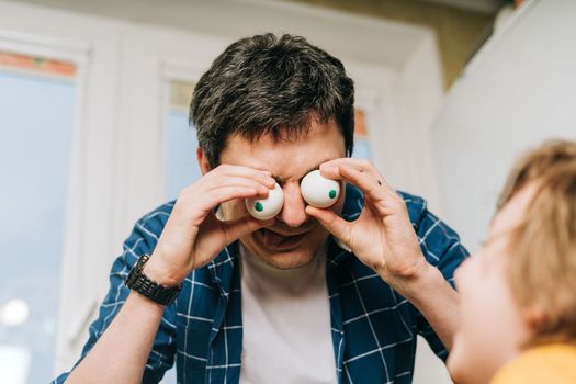 Easter day with kid. Father and son with funny face and eggs eyes, having fun together. Family man and child in a kitchen. Preparing for Easter, creative homemade decoration.